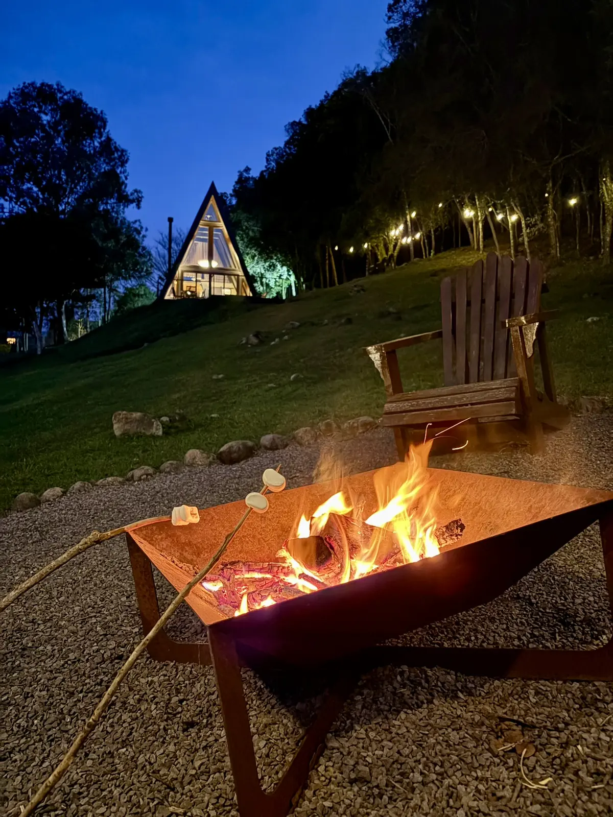 Fogueira acesa à noite com marshmallows, e a cabana iluminada no alto do gramado